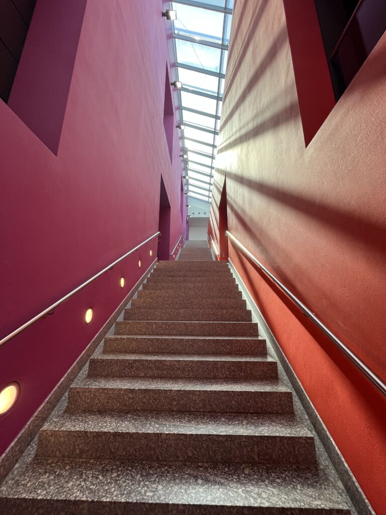 Steile Treppe zwischen roten Wänden mit einfallendem Sonnenlicht durch ein Glasdach.
