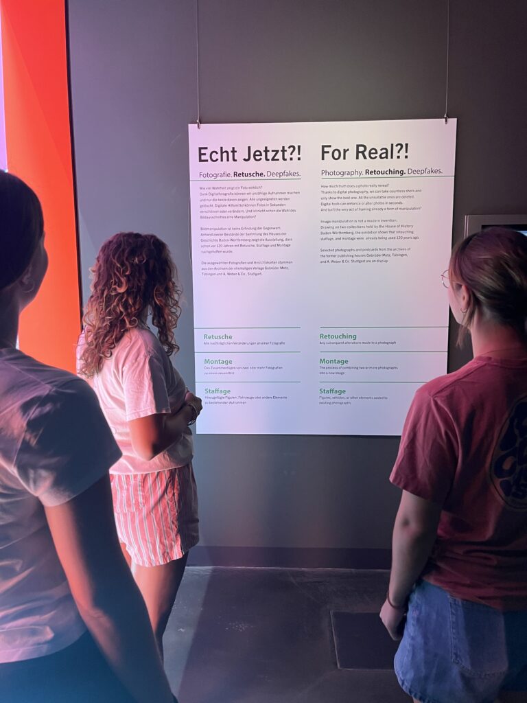 Drei Schülerinnen stehen vor der Einführungstafel zur Ausstellung „Echt jetzt?! Fotografien. Retusche. Deepfakes.“