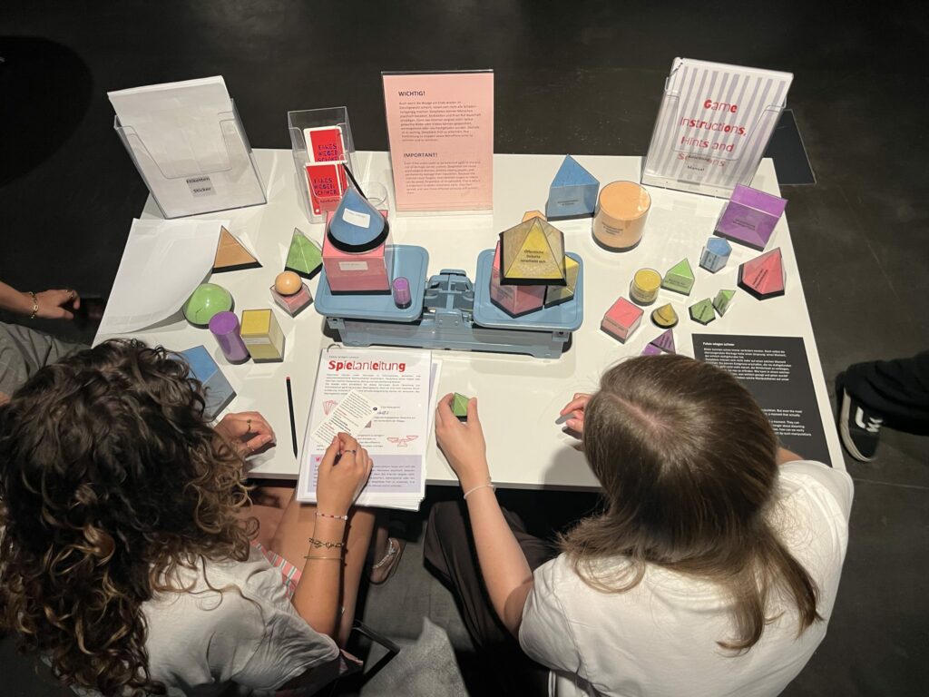 Zwei Personen sitzen an einem Tisch mit bunten geometrischen Formen und Spielmaterial einer interaktiven Ausstellungsstation.