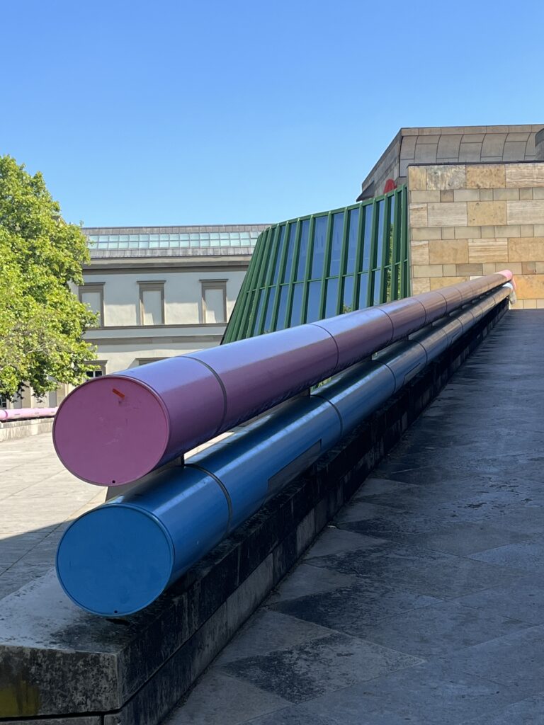Außenansicht der Staatsgalerie Stuttgart mit zwei langen pinken und blauen Rohren vor einer grünen Glasfassade.