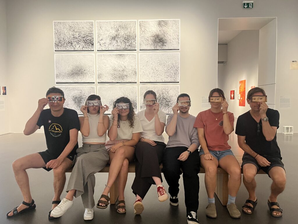 Sieben Personen sitzen nebeneinander auf einer Bank in einem Ausstellungsraum und halten sich kleine Bildkarten vor die Augen.