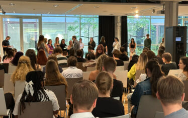 Schülerinnen und Schüler sitzen in der Aula der Johannes-Gutenberg-Schule und verfolgen die feierliche Zeugnisvergabe. Im Vordergrund das Publikum, im Hintergrund werden Absolventinnen und Absolventen auf der Bühne ausgezeichnet.
