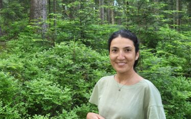 Güllü Kısa steht in einem Wald und lächelt freundlich in die Kamera.