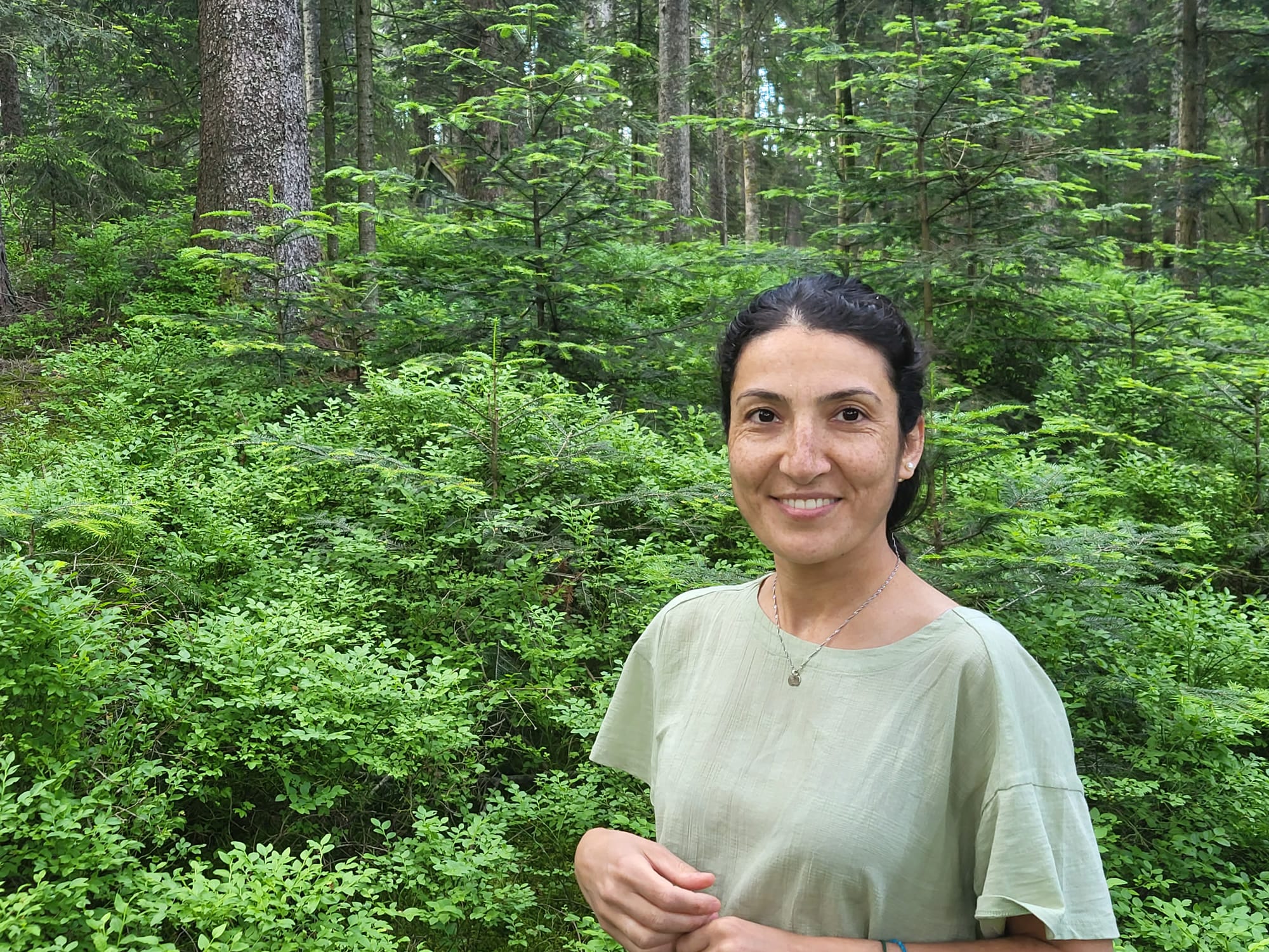Güllü Kısa steht in einem Wald und lächelt freundlich in die Kamera.
