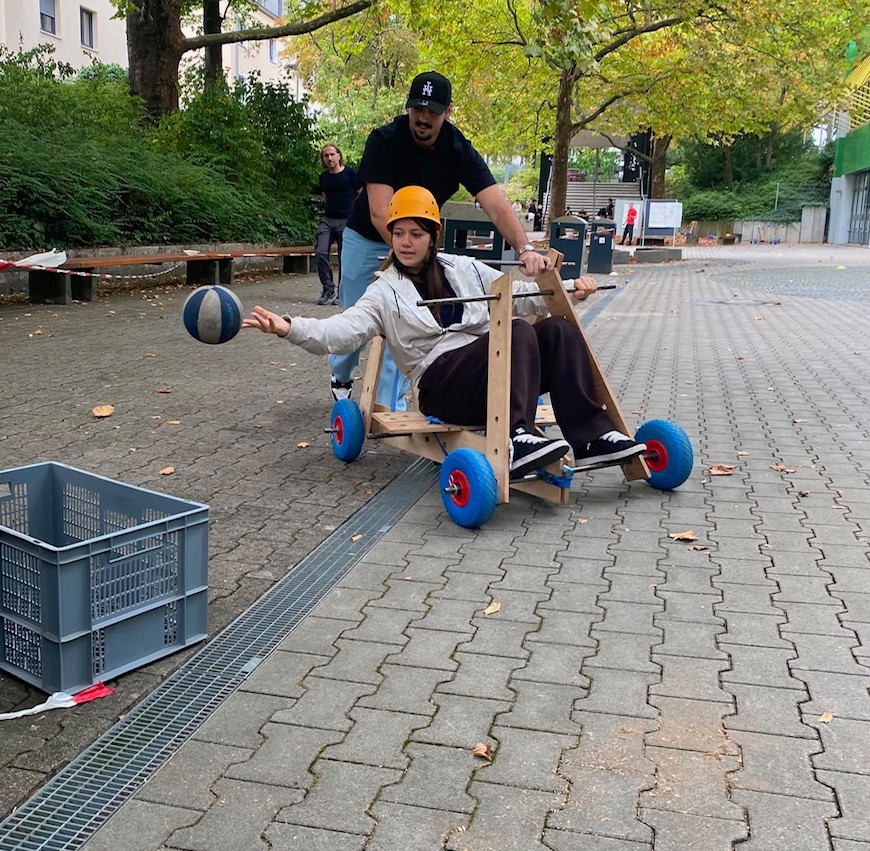 Eine Schülerin sitzt in einer Seifenkiste und versucht während der Fahrt, einen Ball in einen Behälter zu werfen, während ein Mitschüler die Seifenkiste anschiebt.