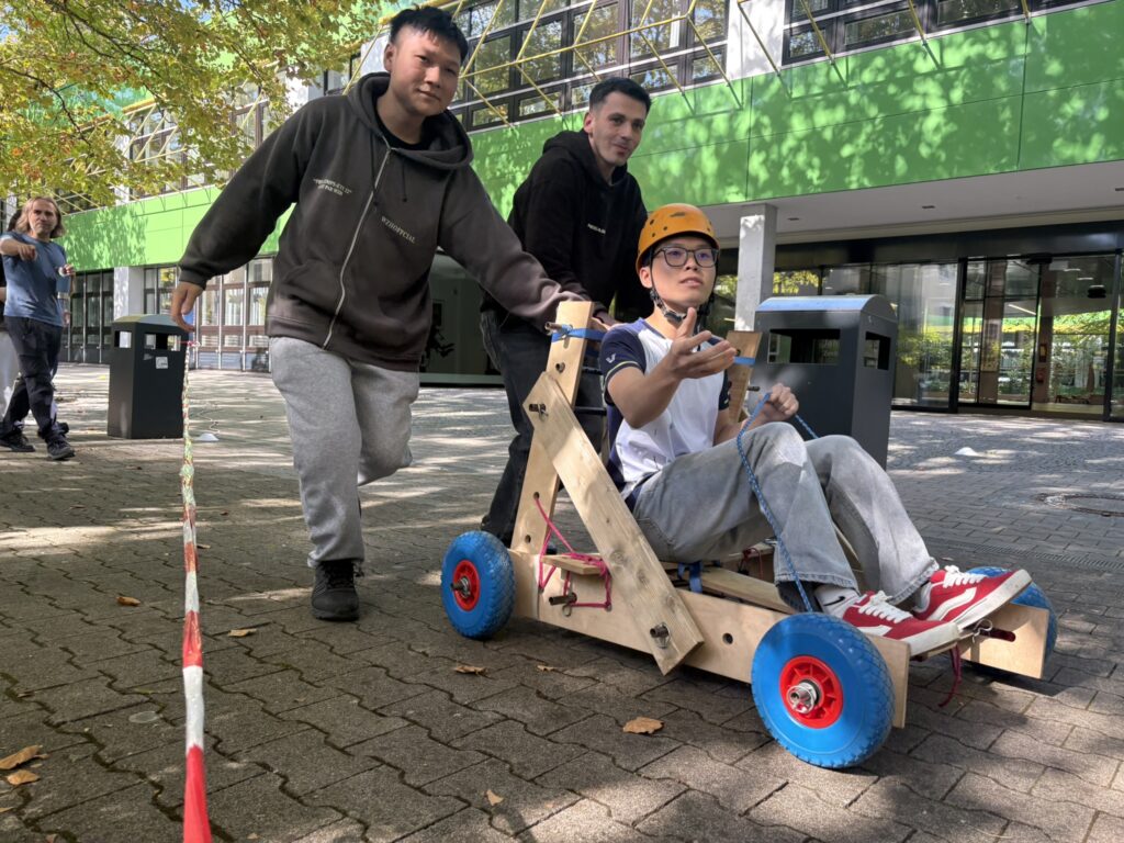 Ein Schüler fährt mit einer selbst gebauten Seifenkiste, während zwei Mitschüler ihn anschieben.