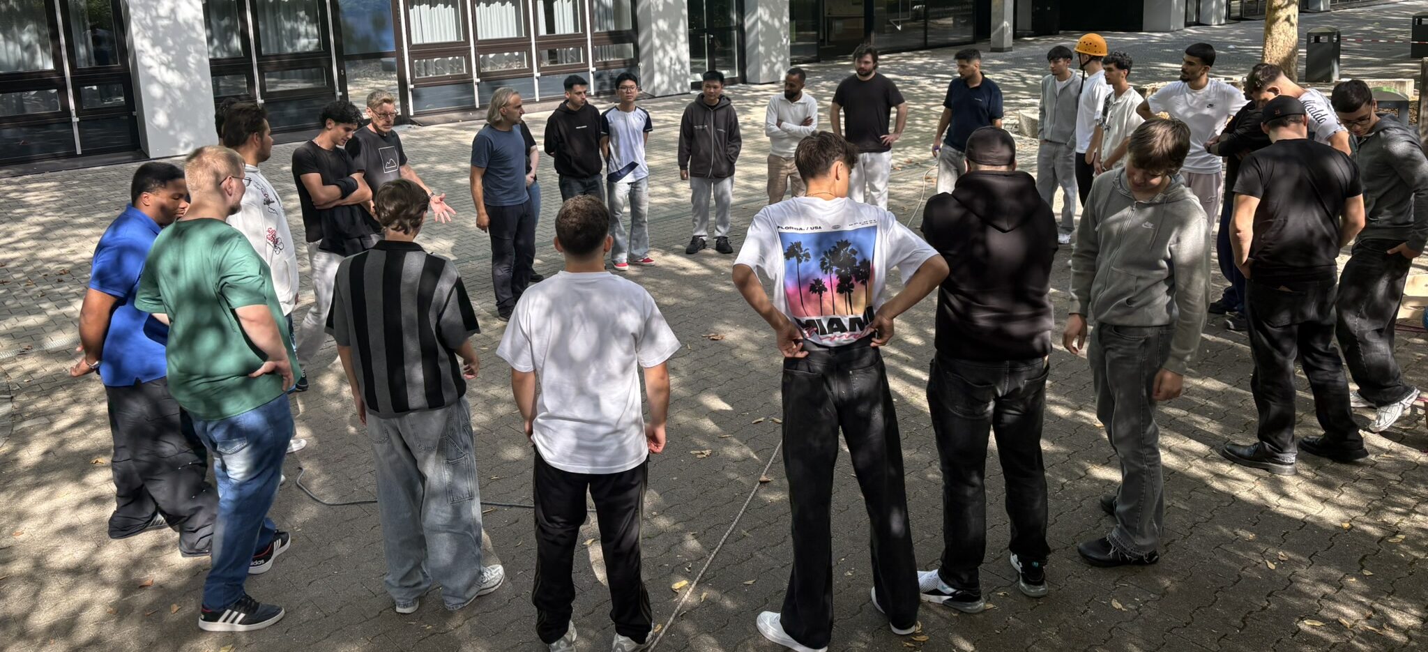 Schüler und Übungsleiter stehen gemeinsam in einem großen Kreis auf dem Schulhof der JGS.