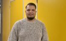 Auf dem Bild ist der neue Lehrer Ilir Markaj vor einer gelben Wand zu sehen. Er steht im Flur der JGS und lächelt freundlich in die Kamera.