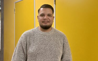Auf dem Bild ist der neue Lehrer Ilir Markaj vor einer gelben Wand zu sehen. Er steht im Flur der JGS und lächelt freundlich in die Kamera.