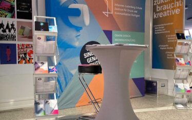 Messestand der Johannes-Gutenberg-Schule mit einer großflächigen Präsentationswand, Informationsbroschüren und einem Stehtisch.