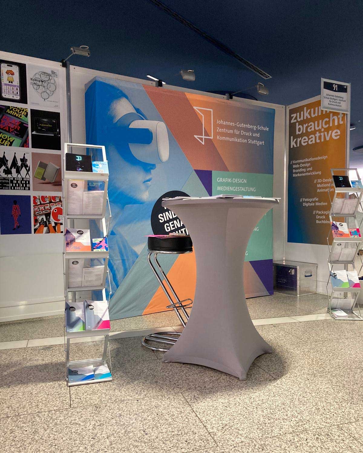 Messestand der Johannes-Gutenberg-Schule mit einer großflächigen Präsentationswand, Informationsbroschüren und einem Stehtisch.