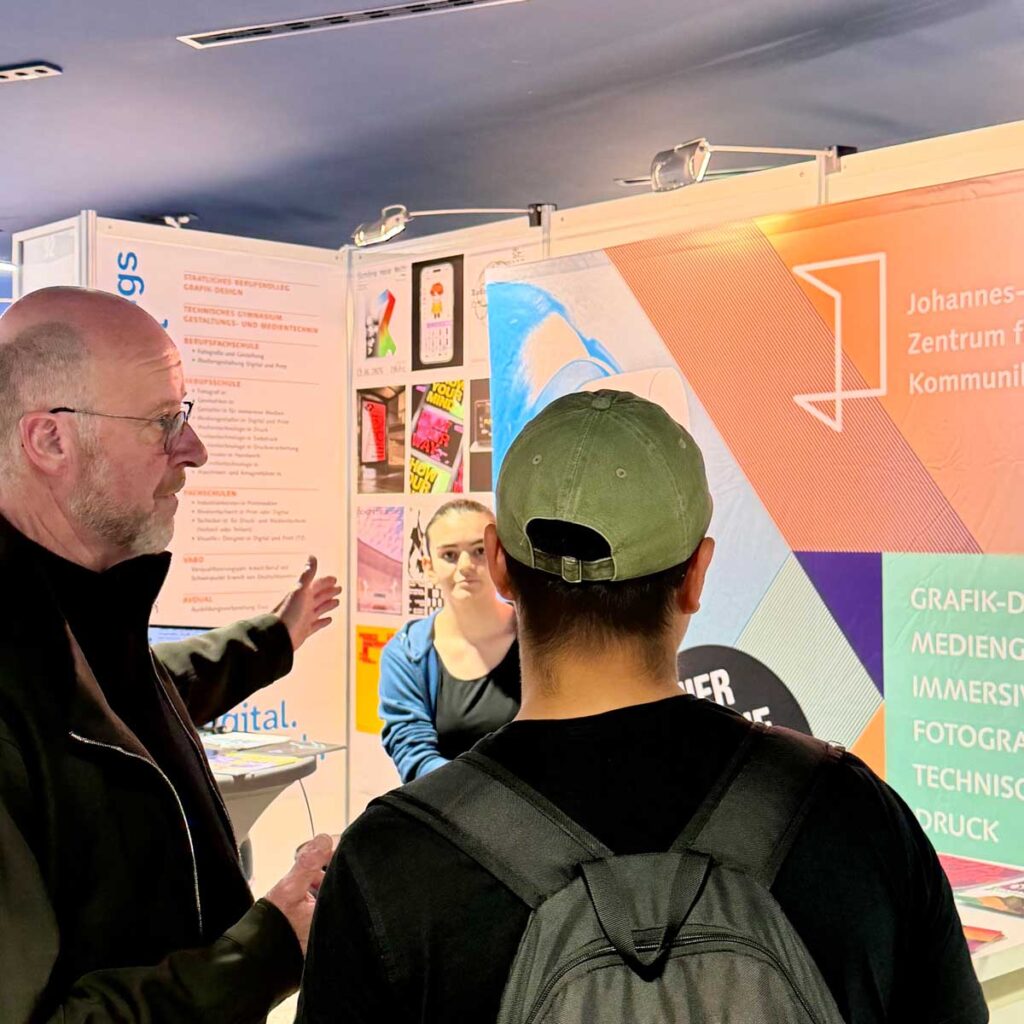 Ein Vertreter der Johannes-Gutenberg-Schule erklärt einem Besucher mit Rucksack die Bildungsangebote am Messestand. Eine weitere Person steht im Hintergrund.