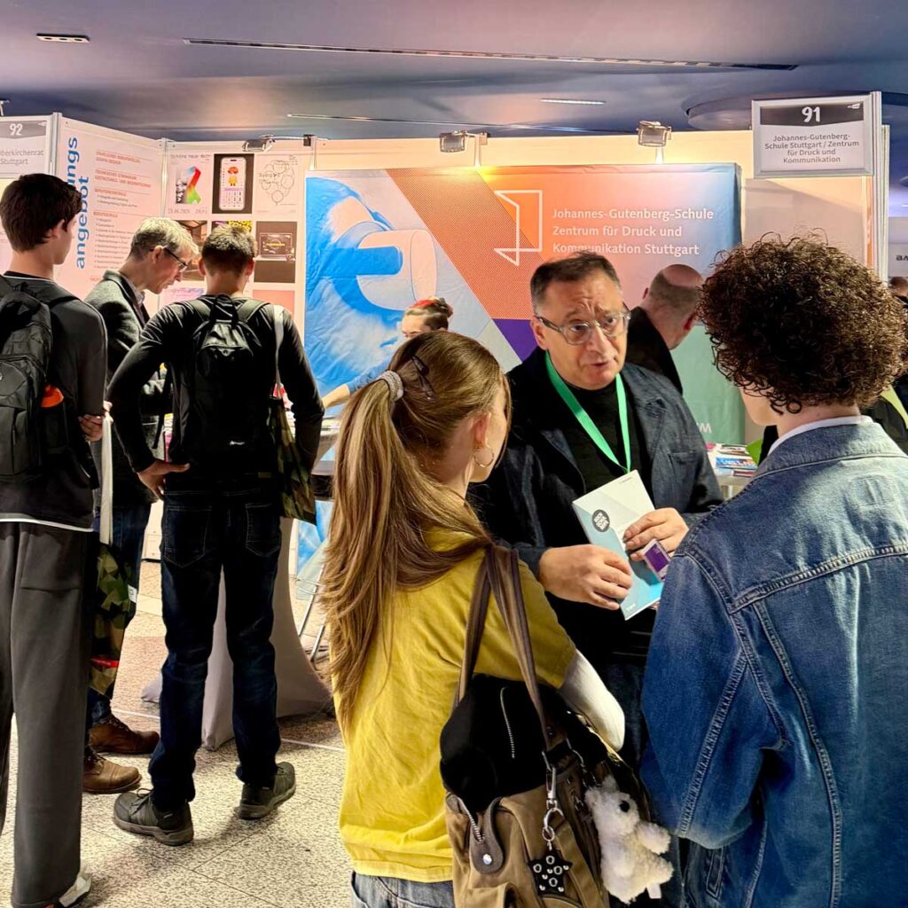 Ein Vertreter der JGS informiert zwei Interessierte am Messestand über die Bildungsangebote der Schule. Im Hintergrund finden weitere Gespräche statt.