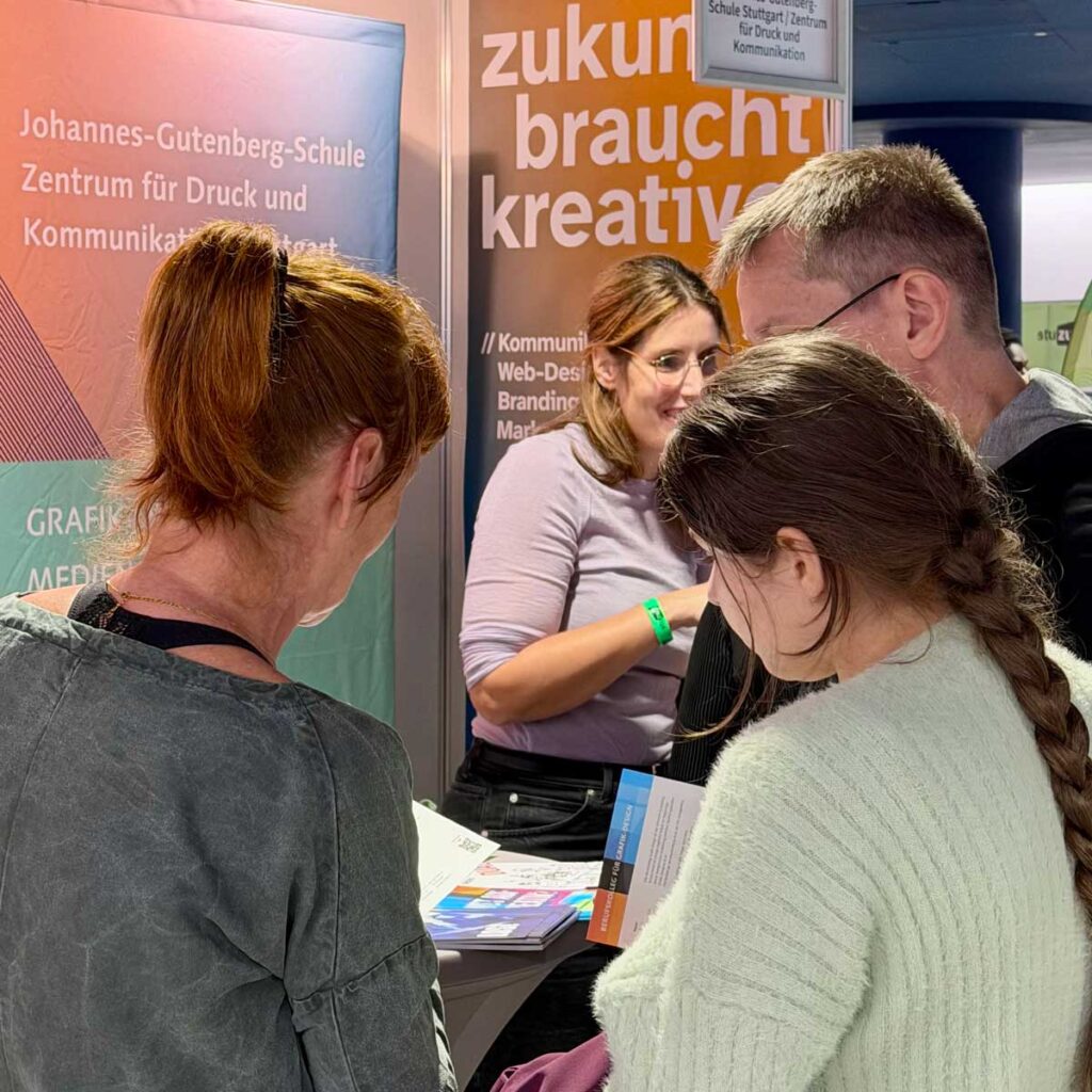 Eine Vertreterin der JGS spricht mit drei Interessierten am Messestand. Auf dem Tisch liegen Informationsbroschüren.
